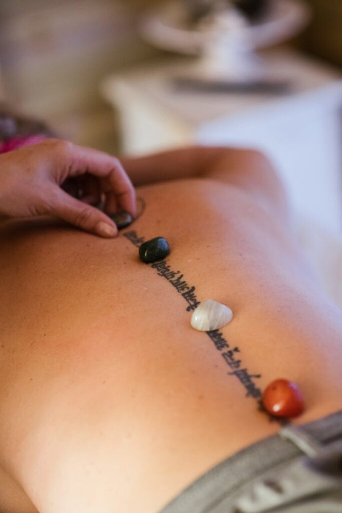 Unrecognizable female patient with tattoo and colorful stones on back getting massage from unrecognizable therapist during massage session in salon on blurred background